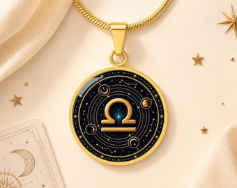 May include: A gold-toned circular pendant necklace featuring the Libra zodiac symbol in the centre. The design includes a dark blue background with concentric circles, celestial stars, and moon phases, all encased in a polished gold frame.