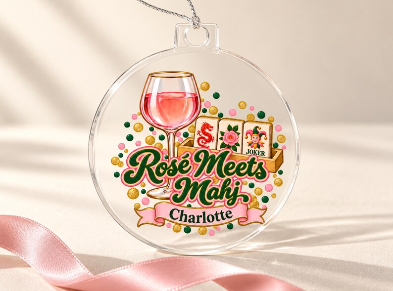 Op de afbeelding: Een doorzichtig, rond, plat ornament met een afbeelding van een wijnglas gevuld met roze vloeistof, drie mahjongtegels en de tekst "Ros&eacute; Meets Mahj" boven een roze banner met de naam "Charlotte" in het zwart, omringd door gekleurde stippen.