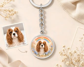 May include: A silver-toned circular keyring featuring a custom portrait of a Cavalier King Charles Spaniel. The dog is depicted against a background of soft clouds, blue flowers, and a rainbow with a small gold heart above its head.