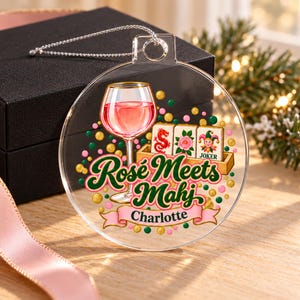 Op de afbeelding: Een doorzichtige, ronde acryl hanger met een afbeelding van een glas ros&eacute;wijn, drie mahjongtegels en de tekst "Ros&eacute; Meets Mahj" boven een roze lint met de naam "Charlotte" in zwarte letters, versierd met kleurrijke confetti stippen.