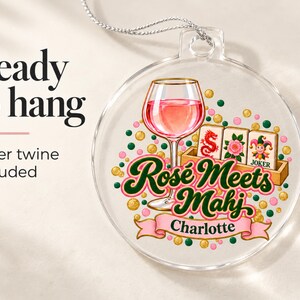 Op de afbeelding: Een doorzichtig rond ornament met een afbeelding van een glas ros&eacute;wijn, drie mahjongtegels en de tekst "Ros&eacute; Meets Mahj" boven een roze banner met de naam "Charlotte". Het ontwerp bevat verspreide roze en groene stippen.