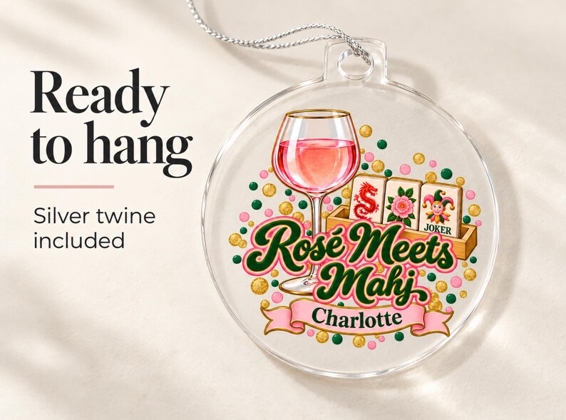 Op de afbeelding: Een doorzichtig rond ornament met een afbeelding van een glas ros&eacute;wijn, drie mahjongtegels en de tekst "Ros&eacute; Meets Mahj" boven een roze banner met de naam "Charlotte". Het ontwerp bevat verspreide roze en groene stippen.