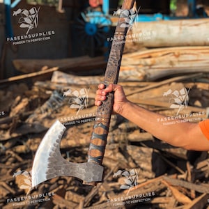 Può includere: Un'ascia artigianale con manico in legno e lama color argento. Il manico è avvolto in pelle nera e presenta incisioni decorative. Il testo "FASEEHSUPPLIES FORGED TO PERFECTION" è visibile.