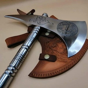 May include: A polished steel axe with a decorative lion's head emblem and etched designs. The axe features a silver handle and is paired with a brown leather sheath. The sheath has a lion's head embossed design and is secured with leather straps.