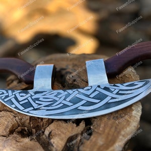 Könnte beinhalten: Eine silberne Axt mit einem Holzstiel. Die Axtklinge hat ein keltisches Knotendesign und ist gebogen. Die Axt liegt auf einem Stück Holz.