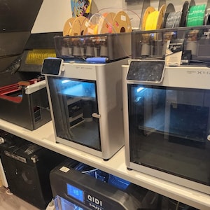 Peut inclure: Deux imprimantes 3D argentées Bambu Lab X1-Carbon avec unités AMS sur le dessus, dotées de portes vitrées et d'écrans tactiles. À gauche se trouve une découpeuse laser noire avec un tuyau d'aération, et en dessous une imprimante QIDI X-Max.