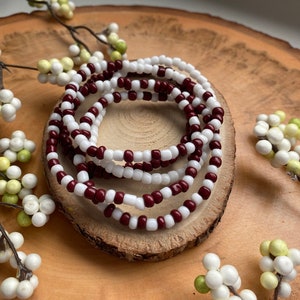 May include: A set of two beaded bracelets, one white and one maroon, are stacked on top of each other. The bracelets are made of small, round beads.