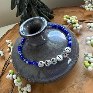 May include: A blue and silver beaded bracelet with the word "RESIST" spelled out in silver beads.
