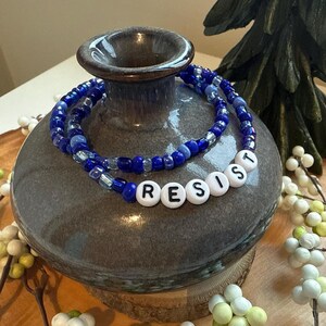 May include: A blue and white beaded necklace with the word "RESIST" spelled out in white beads.