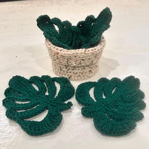 Puede incluir: Juego de ganchillo hecho a mano con una pequeña cesta beige moteada con hojas de ganchillo verde oscuro en el interior. Dos posavasos de hoja de monstera de ganchillo verde oscuro están delante de la cesta. El juego está sobre una superficie blanca.