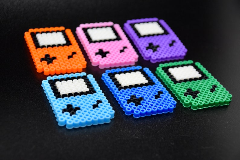 May include: Six colorful pixel art Game Boy style keychains. The keychains are made of plastic beads and feature different colors, including orange, pink, purple, blue, green, and light blue.