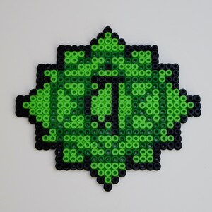 Op de afbeelding: Een groen en zwart pixel art ontwerp, dat lijkt op een gestileerd oog, gemaakt met kleine, ronde kralen. De oogvorm is omgeven door een zwarte rand en het midden heeft de zwarte letter "I".