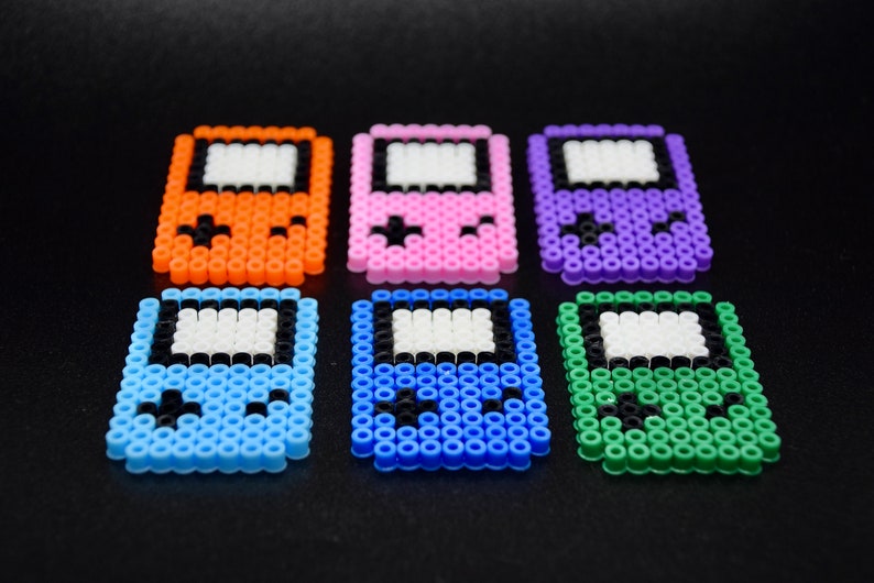 May include: Six colorful pixel art game console keychains. The keychains are made of plastic beads and feature different colors, including orange, pink, purple, blue, and green.