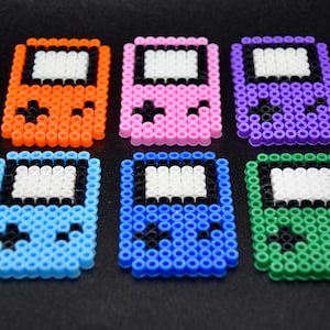 May include: Six colorful pixel art game consoles made with perler beads. The consoles are orange, pink, purple, blue, green, and light blue.