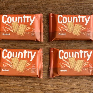 May include: Four rectangular snack packages labelled Country Pretzel in white text on an orange background. Each package features an illustration of two rectangular crackers and wheat stalks, with a small vegan symbol and an 8x count indicator.