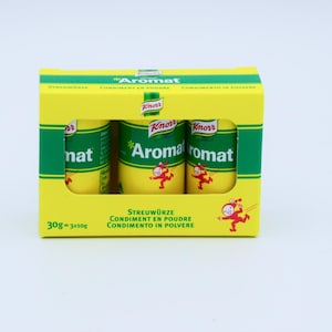 Könnte beinhalten: Eine gelbe Schachtel mit drei kleinen, zylindrischen Gewürzbehältern mit der Aufschrift "Aromat" in Grün und Weiß. Jeder Behälter hat eine Cartoonfigur. Die Schachtel enthält auch Text in mehreren Sprachen, darunter "Condiment en poudre".