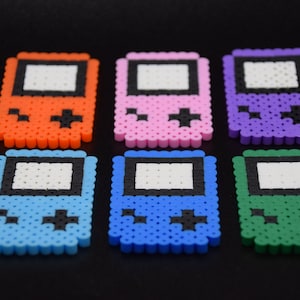 May include: Six colorful pixel art game console keychains. The keychains are made of plastic beads and feature a variety of colors, including orange, pink, purple, blue, light blue, and green.