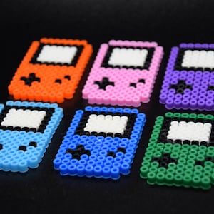 May include: Six colorful pixel art game consoles made with perler beads. The consoles are in orange, pink, purple, blue, green, and light blue.