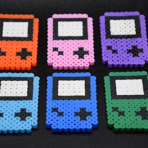 May include: Six colorful pixel art Game Boy keychain charms. The charms are made of plastic beads and feature a classic Game Boy design with a black screen and buttons. The charms are orange, pink, purple, light blue, blue, and green.