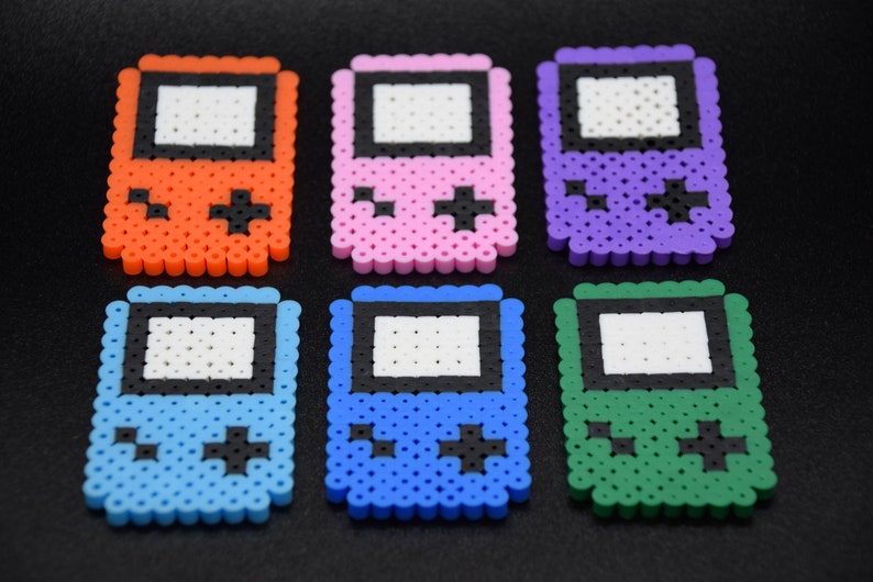 May include: Six colorful pixel art Game Boy keychain charms. The charms are made of plastic beads and feature a classic Game Boy design with a black screen and buttons. The charms are orange, pink, purple, light blue, blue, and green.