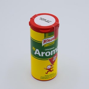 Könnte beinhalten: Ein gelber und grüner zylindrischer Behälter mit einem roten Deckel. Der Behälter trägt den Text "Knorr* Original Aromat". Der Behälter hat auch ein kleines Bild einer Person, die eine Flagge mit einem weißen Kreuz auf rotem Hintergrund hält. Der Text "Streuwürze Condiment en poudre Condimento in polvere" ist auf der Unterseite des Behälters aufgedruckt.