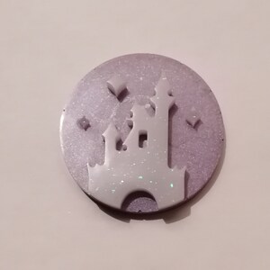 May include: A white castle silhouette with glitter accents on a purple and white oval background.