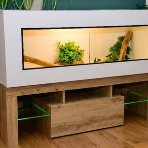 May include: A white rectangular reptile enclosure with a clear glass front panel, featuring internal artificial green plants and wooden branches. The enclosure sits on a light brown wood-grain stand with open shelving and glowing green glass shelves.