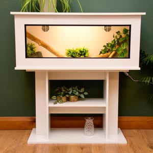 May include: A white reptile vivarium with a glass front, mounted on a matching stand. Inside, there's a branch, greenery, and warm lighting. The stand has shelves with plants and a decorative glass vase.