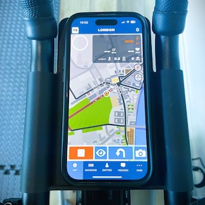 May include: A smartphone displaying a map app with the word "LONDON" on the screen. The map shows a zoomed-in view of a city with streets and buildings. The app has buttons for "MAP", "DASHBOARD", "ZWIFTERS", "MESSAGES", and "MORE".