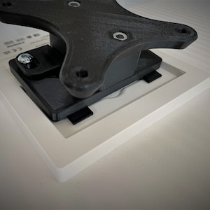 May include: A black plastic wall mount with a square white frame attached to it. The mount has a screw in the top left corner.