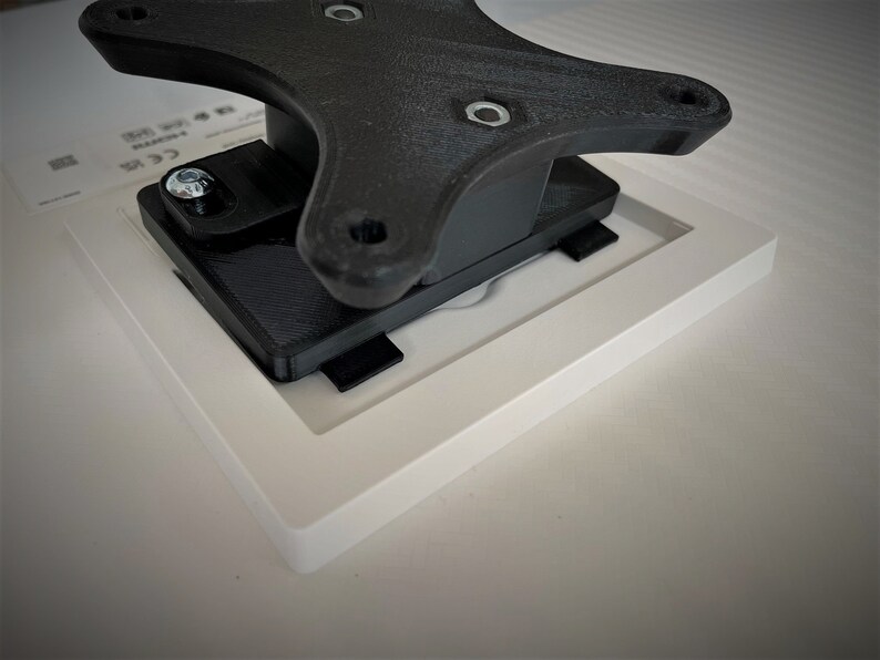 May include: A black plastic wall mount with a square white frame attached to it. The mount has a screw in the top left corner.
