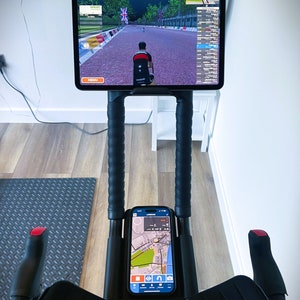 May include: A black stationary exercise bike with a tablet mounted on the handlebars. The tablet is displaying a cycling app with a virtual race. A smartphone is also mounted on the handlebars, displaying a map.