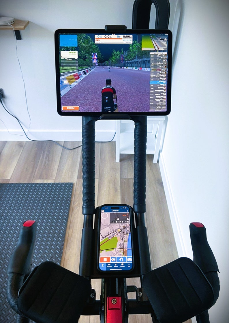 May include: A black stationary exercise bike with a tablet mounted on the handlebars. The tablet is displaying a cycling app with a virtual race. A smartphone is also mounted on the handlebars, displaying a map.