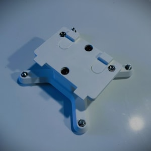 May include: White plastic mounting bracket with four screw holes and a rectangular opening. The bracket has a textured surface and is designed for mounting a device.