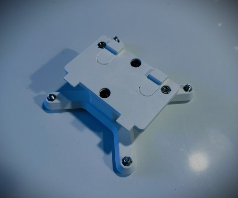 May include: White plastic mounting bracket with four screw holes and a rectangular opening. The bracket has a textured surface and is designed for mounting a device.