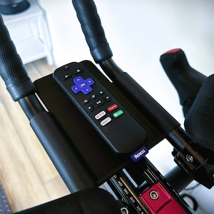 May include: A black remote control with a purple Roku logo is mounted on a black handlebar. The remote has buttons for navigation, playback, and streaming services like Netflix and YouTube.