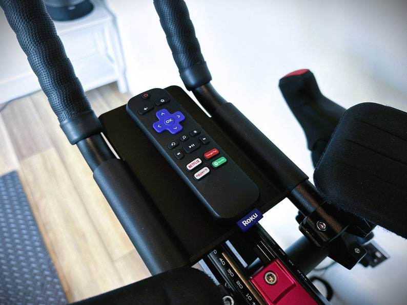 May include: A black remote control with a purple Roku logo is mounted on a black handlebar. The remote has buttons for navigation, playback, and streaming services like Netflix and YouTube.