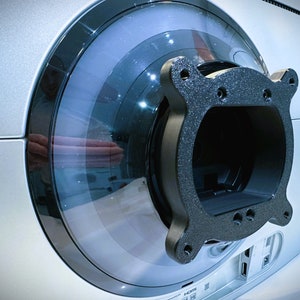 May include: Close-up of a silver and black electronic device. The device has a circular design with a transparent, blue-tinted section. A black, textured bracket is attached to the device, featuring multiple screw holes.