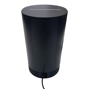 Puede incluir: Un altavoz inteligente cilíndrico negro con un cable de alimentación enchufado. El altavoz tiene una pequeña luz en la parte superior y un micrófono en la parte delantera.