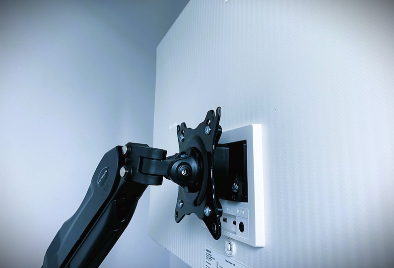 May include: A black monitor arm mounted to a white wall plate. The arm is extended and angled towards the left. The wall plate has a white trim and a black electrical outlet.