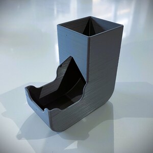 May include: A gray 3D printed dice tower with a square top and a curved bottom. The tower has a slot on the side for rolling dice.