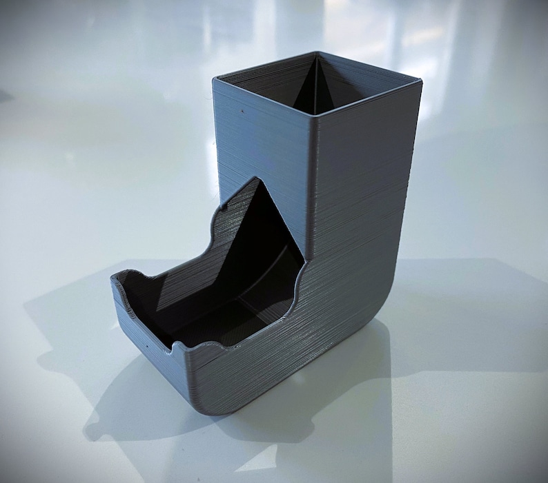 May include: A gray 3D printed dice tower with a square top and a curved bottom. The tower has a slot on the side for rolling dice.