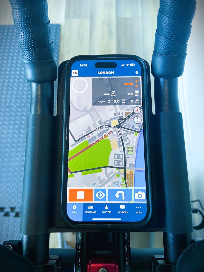 May include: A smartphone displaying a map app with the word "LONDON" on the screen. The map shows a zoomed-in view of a city with streets and buildings. The app has buttons for "MAP", "DASHBOARD", "ZWIFTERS", "MESSAGES", and "MORE".
