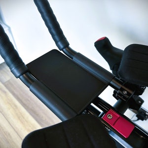 May include: Black exercise bike handlebars with padded grips and a black padded armrest. The handlebars have a red button with the text '+00'.