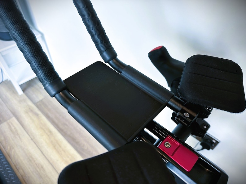 May include: Black exercise bike handlebars with padded grips and a black padded armrest. The handlebars have a red button with the text '+00'.