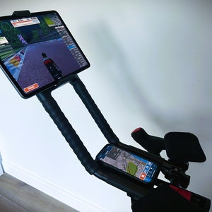 May include: A black exercise bike with a tablet mounted on the handlebars. The tablet is displaying a cycling app with a virtual route. A smartphone is also mounted on the handlebars, displaying a map.
