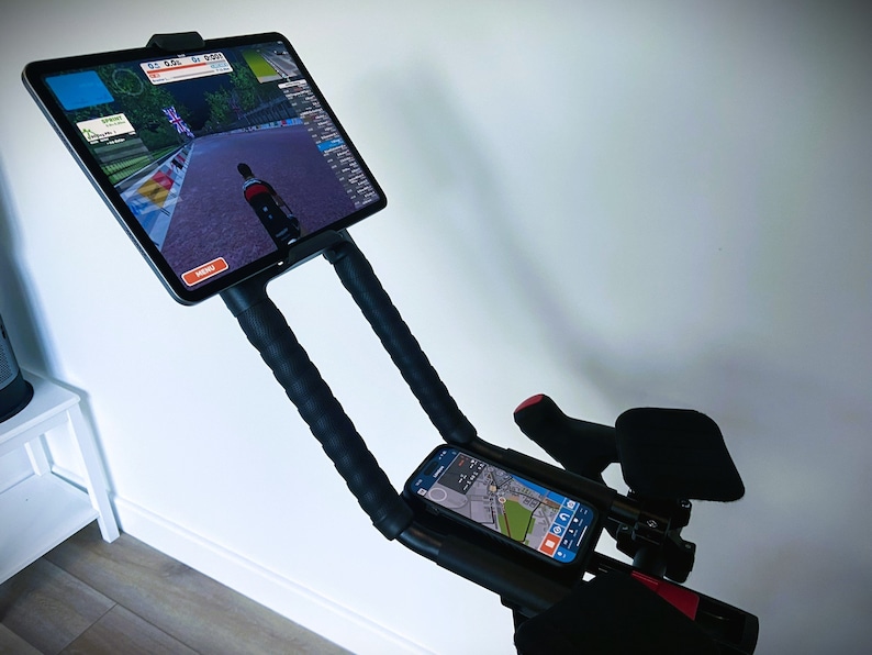 May include: A black exercise bike with a tablet mounted on the handlebars. The tablet is displaying a cycling app with a virtual route. A smartphone is also mounted on the handlebars, displaying a map.