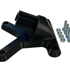 Op de afbeelding: Een zwarte 3D-geprinte beugel met een rechthoekig bovendeel en schuine steunarmen. De beugel heeft verschillende montagegaten en wordt vergezeld door een set zilveren bouten en moeren, waarschijnlijk voor montage.