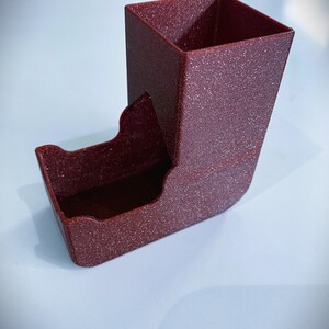 May include: A red dice tower with a textured finish. The tower has a square top and a curved bottom.