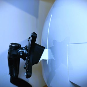 Puede incluir: Un soporte de TV de metal negro con un brazo giratorio y una cubierta de plástico blanco. El soporte está fijado a una pared blanca.